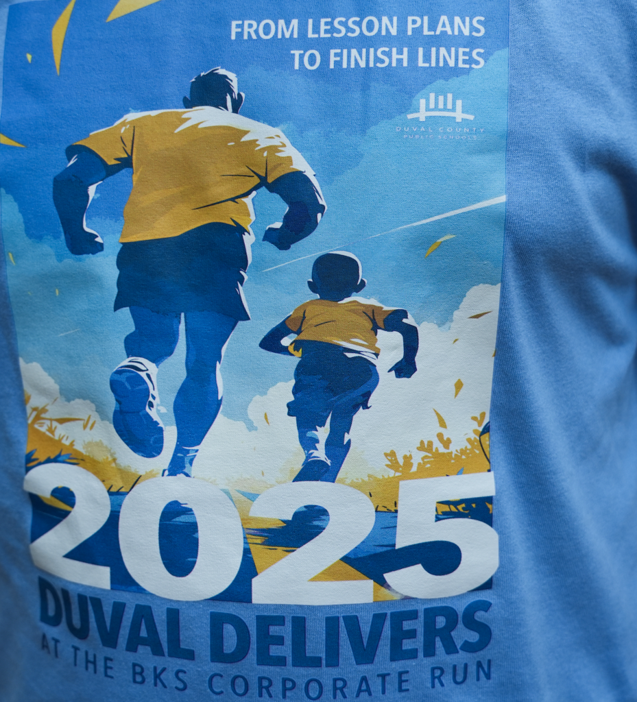 Close-up of the 2025 Team Duval race shirt featuring two runners — an adult and a child — with the slogan “From Lesson Plans to Finish Lines” and the words “Duval Delivers at the BKS Corporate Run.”