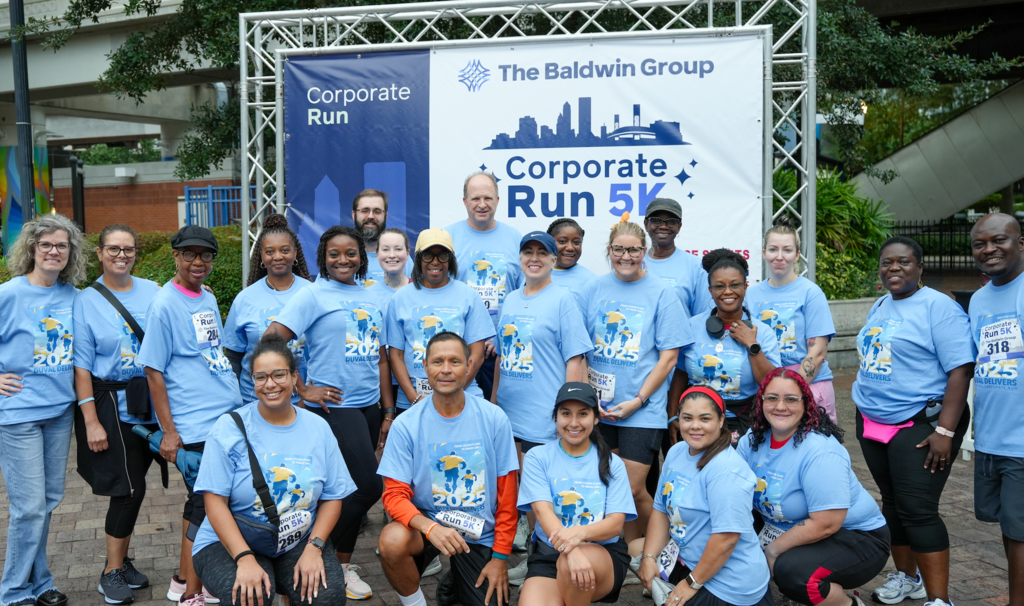 Team Duval participants pose in front of the Baldwin Group Corporate Run 5K banner, all wearing the official “Duval Delivers 2025” shirts. The group stands proudly together, celebrating their participation and teamwork at the Jacksonville event.