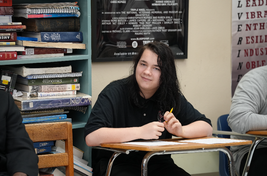 A Westside High School student smiles while participating in the Veterans Day Road Show discussion, surrounded by history books and classroom posters.