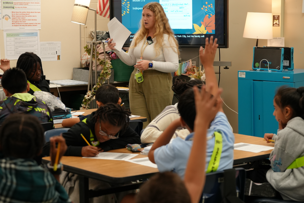 Edina Besic addressing her fifth-grade class as students raise their hands to answer a question, surrounded by colorful classroom decorations.