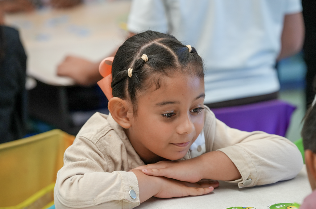 A first-grade student leans forward on her desk, smiling thoughtfully as she listens during a classroom activity.
