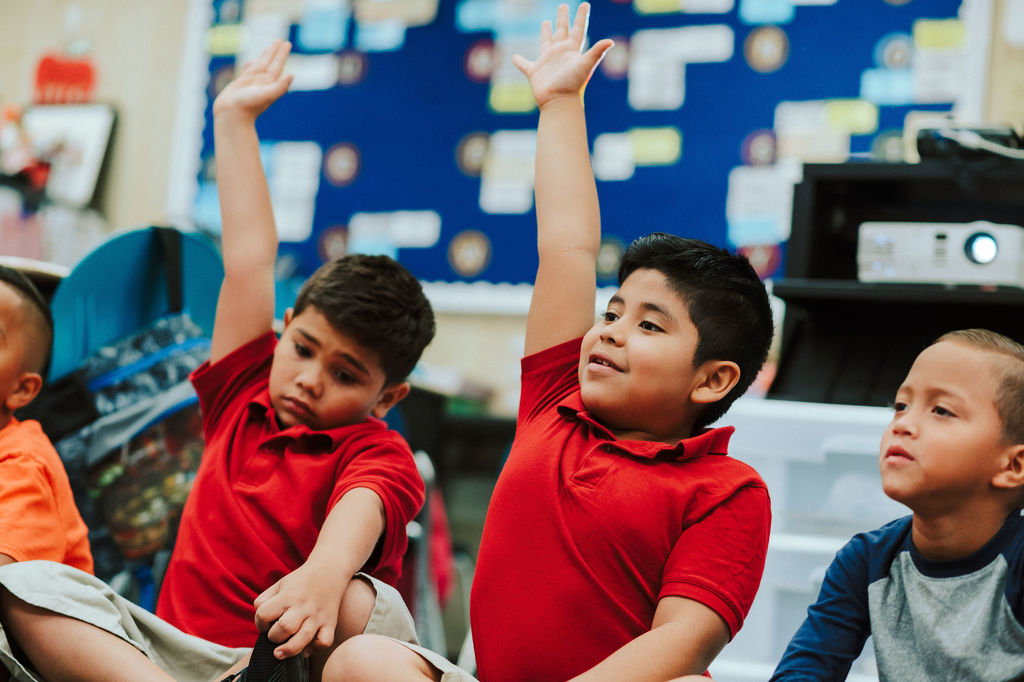 A photo of three elementary-school students sitting on the classroom floor.  Two of students have their hands raised high. 