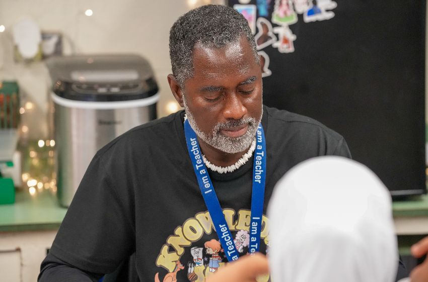 Vincent Taylor smiles while interacting with a student during class. He wears glasses and a blue Boys & Girls Club T-shirt, and the student beside him is smiling as well.