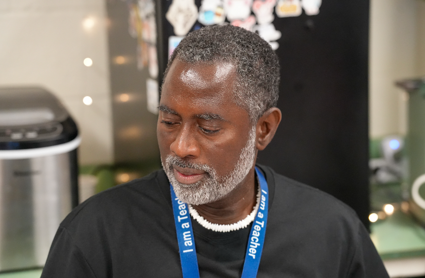 Vincent Taylor smiles while interacting with a student during class. He wears glasses and a blue Boys & Girls Club T-shirt, and the student beside him is smiling as well.