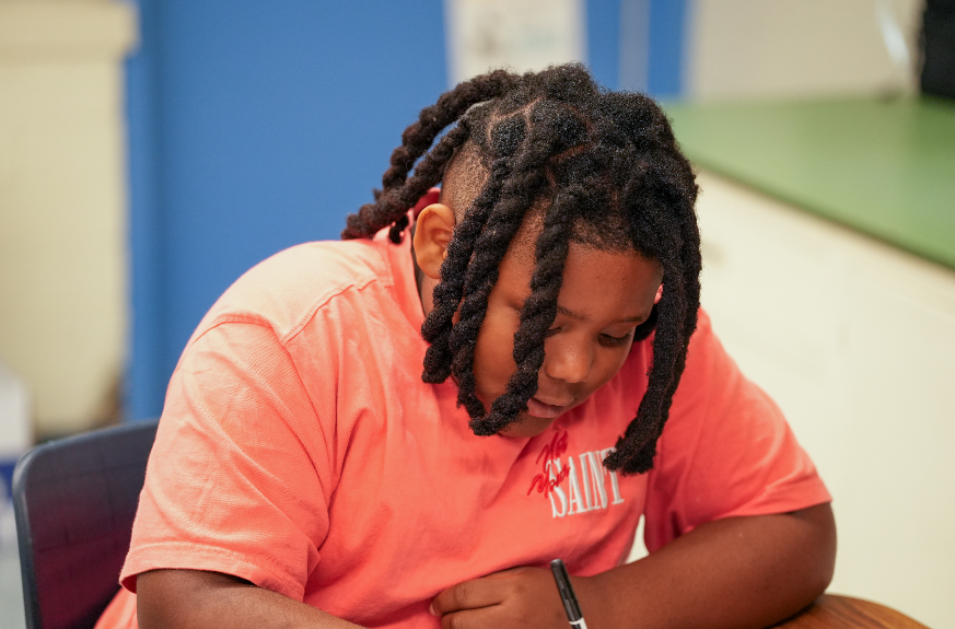 A student in a coral-colored shirt leans over a desk while writing on paper, concentrating on their work inside a classroom.