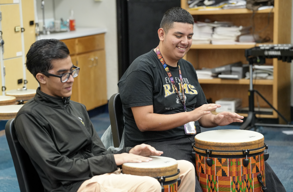 A student taps a hand drum while sitting with classmates during a music lesson.