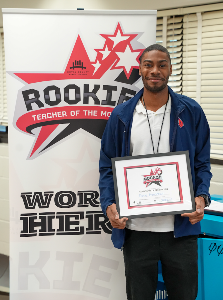 Davie Henderson, a fifth-grade teacher at Crystal Springs Elementary, stands smiling in a classroom holding his Rookie Teacher of the Month certificate, with a Rookie Teacher of the Month banner and football-themed posters visible behind him.