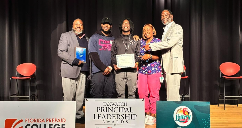 Principal Hall stands with student Owen and family members on stage as Owen holds a certificate during a Florida TaxWatch Principal Leadership Award ceremony.