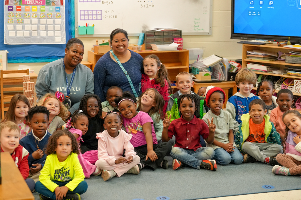 Teacher Erika Cunamay and classroom staff member sit with pre-primary Montessori students gathered on a classroom rug, smiling together at John E. Ford PK-8 Montessori.