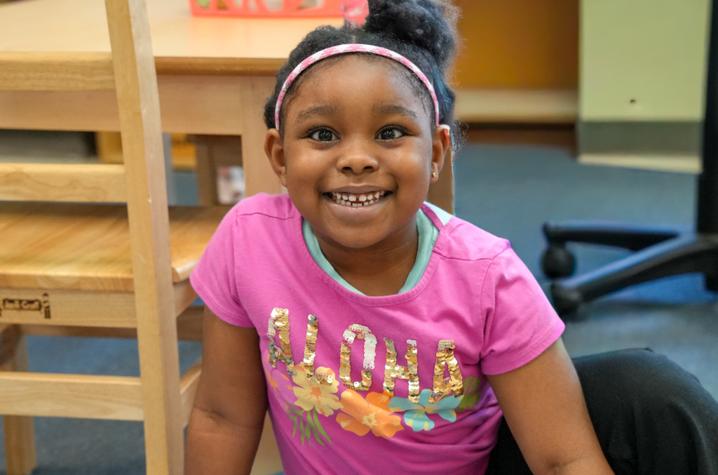 Student in a pink shirt smiling while holding Montessori learning materials in the classroom.