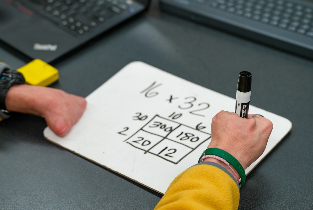 A close-up shows Benton Jones writing a math equation and grid diagram on a small whiteboard during instruction.