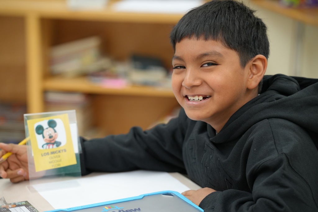 An elementary student smiles while working at a desk inside a classroom.