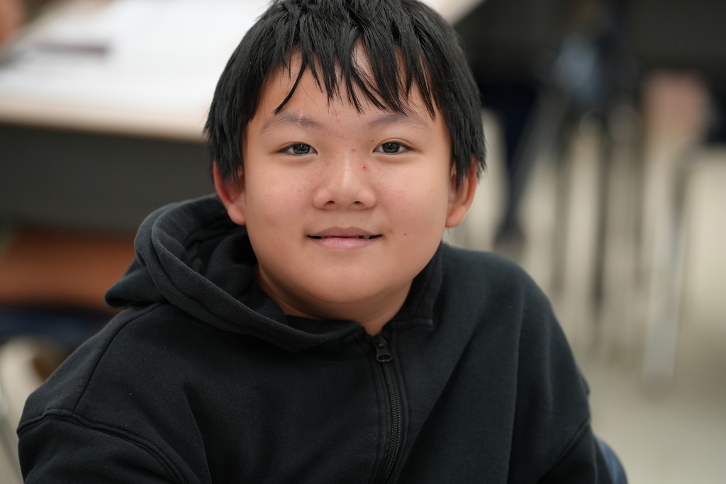 A smiling elementary student looks directly at the camera inside a classroom setting.