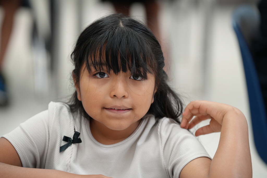 An elementary student with braided hair looks toward the camera with a neutral expression inside a classroom.