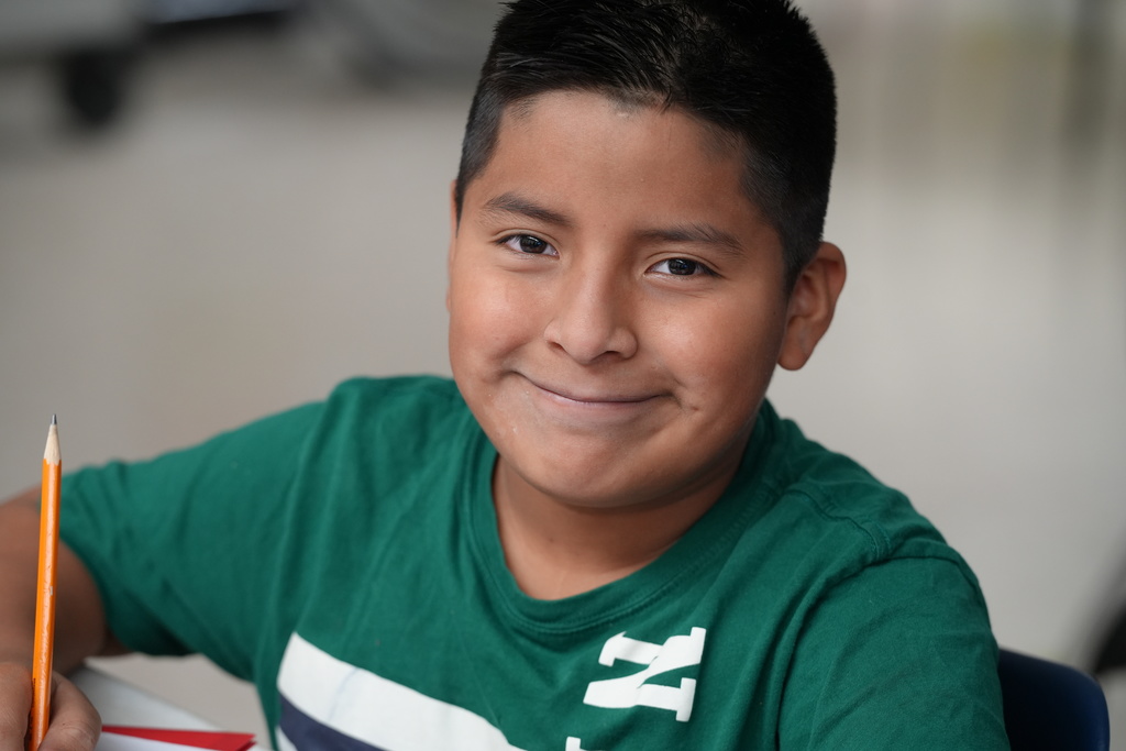 A smiling elementary student wearing a green shirt looks at the camera inside a classroom.