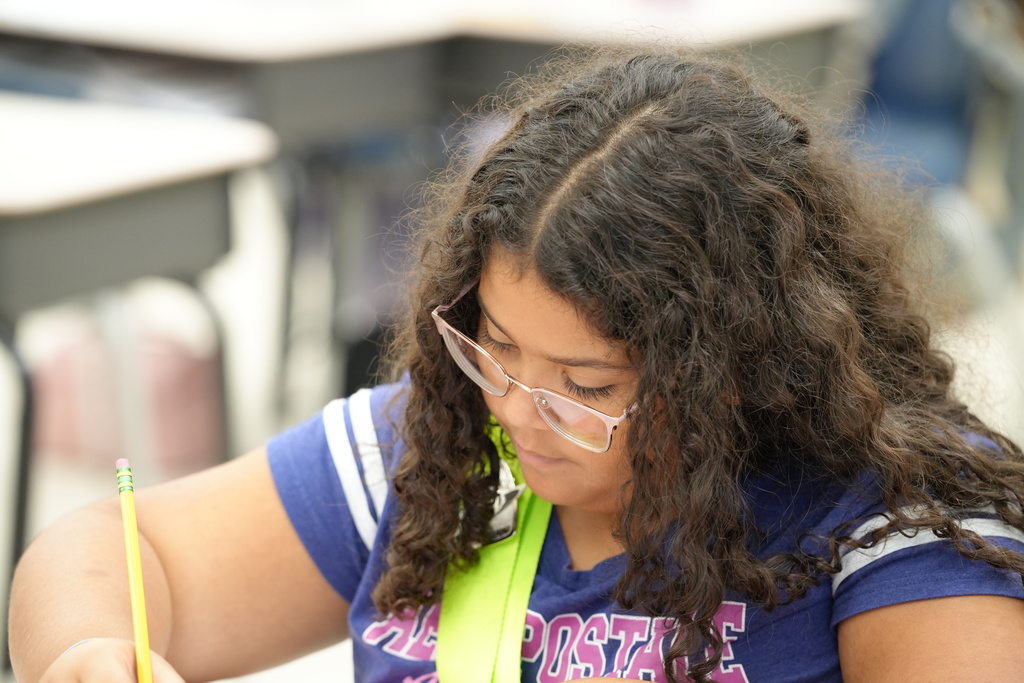 A student focuses on schoolwork at a desk with classroom materials in the background.