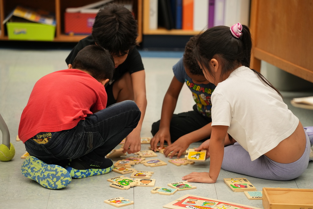 Three elementary students sit on the classroom floor working together on a hands-on learning activity with wooden puzzle pieces spread out in front of them.