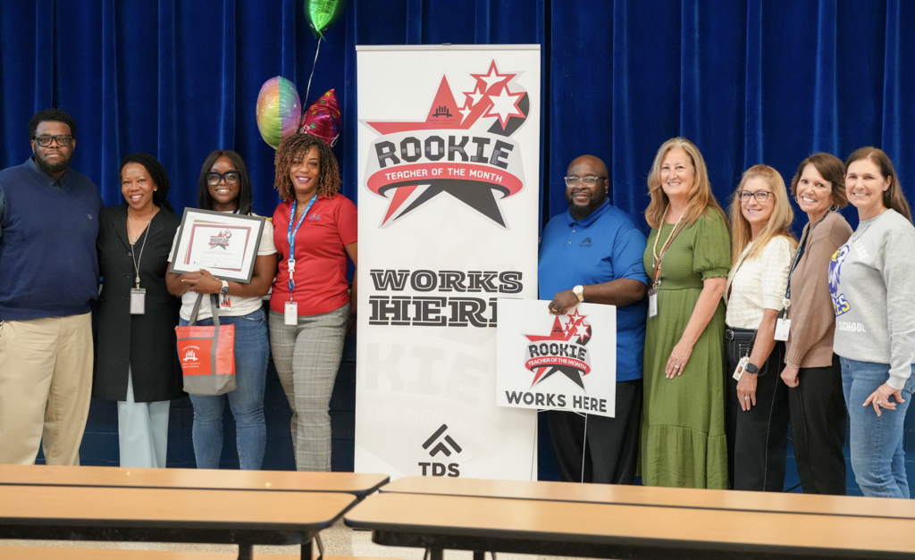 Mrs. Dankeshia Ivester stands with administrators and staff on a stage next to a “Rookie Works Here” banner while holding her award sign and certificate.
