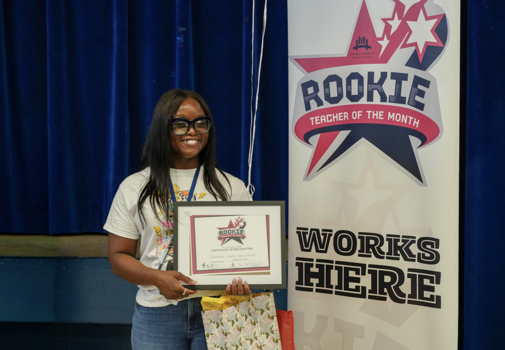 Mrs. Dankeshia Ivester poses with school and district staff beside a “Rookie Works Here” banner, holding a recognition sign during a celebration at Normandy Village Elementary School.