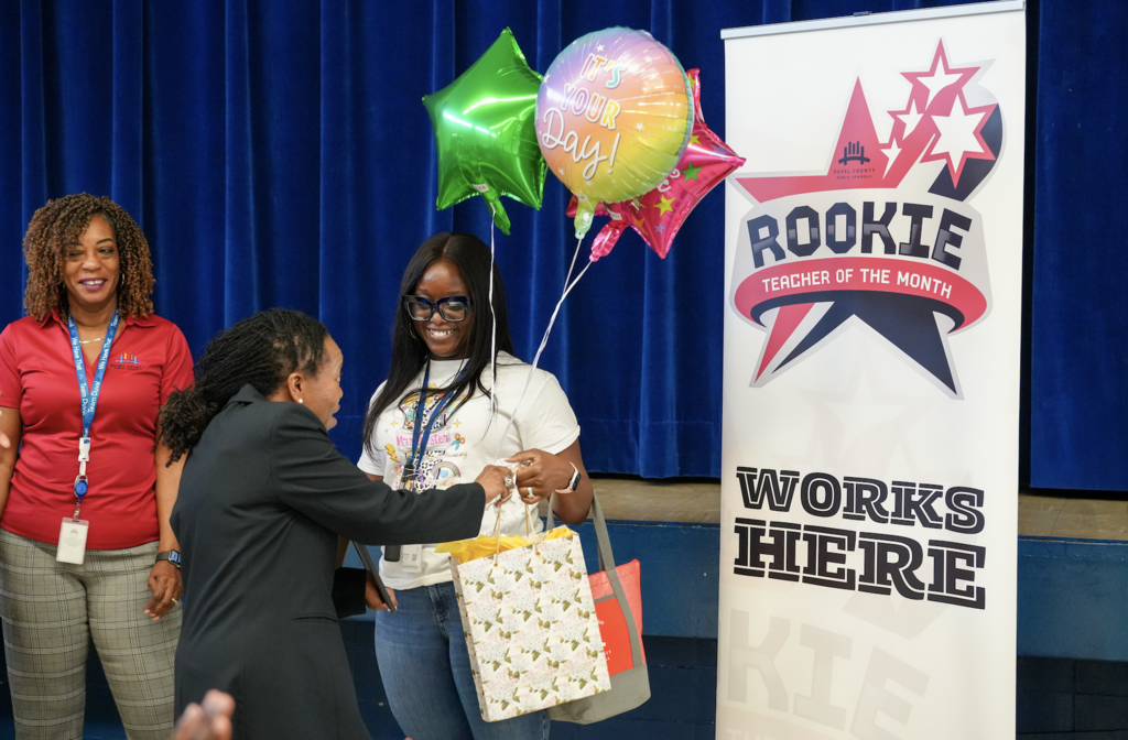 Mrs. Dankeshia Ivester stands with colleagues while holding her Rookie Counselor of the Month certificate in front of a blue stage curtain with balloons in the background.