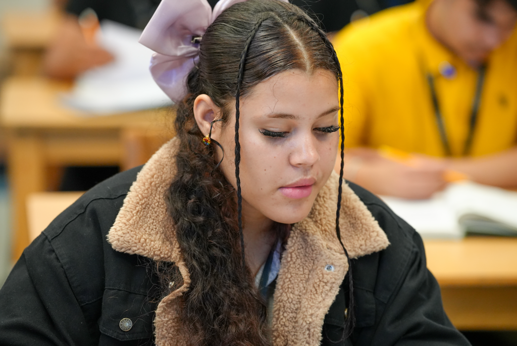 A female student with braided hair and a pink bow concentrates while reading and writing at her desk in a classroom.