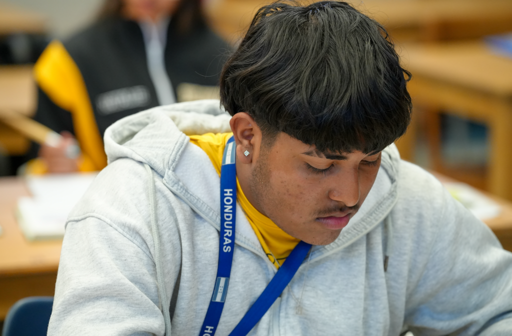 A male student wearing a gray hoodie and a Honduras lanyard looks down as he focuses on his classwork.