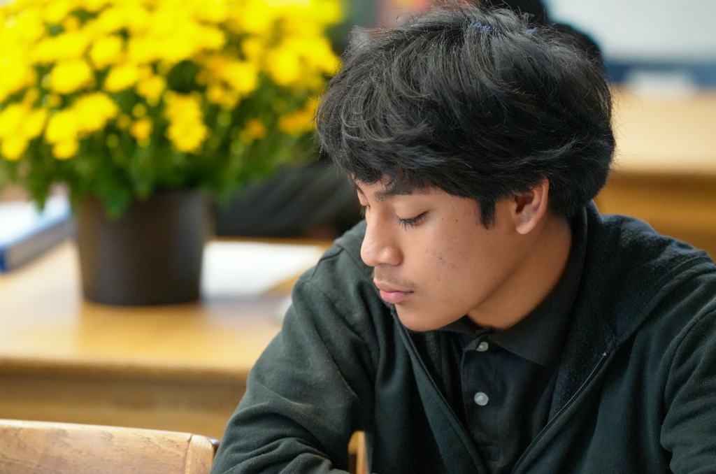 A male student in a black polo leans over his desk, concentrating on his assignment with a yellow potted plant in the background.