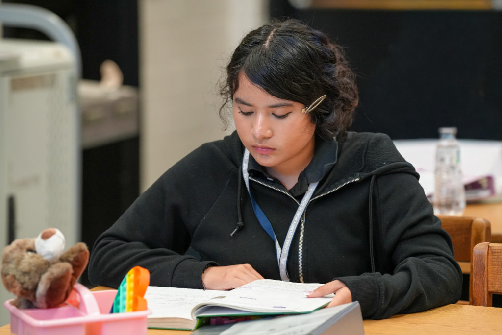 A female student in a black hoodie reads intently from an open book at her desk, with classroom supplies nearby.