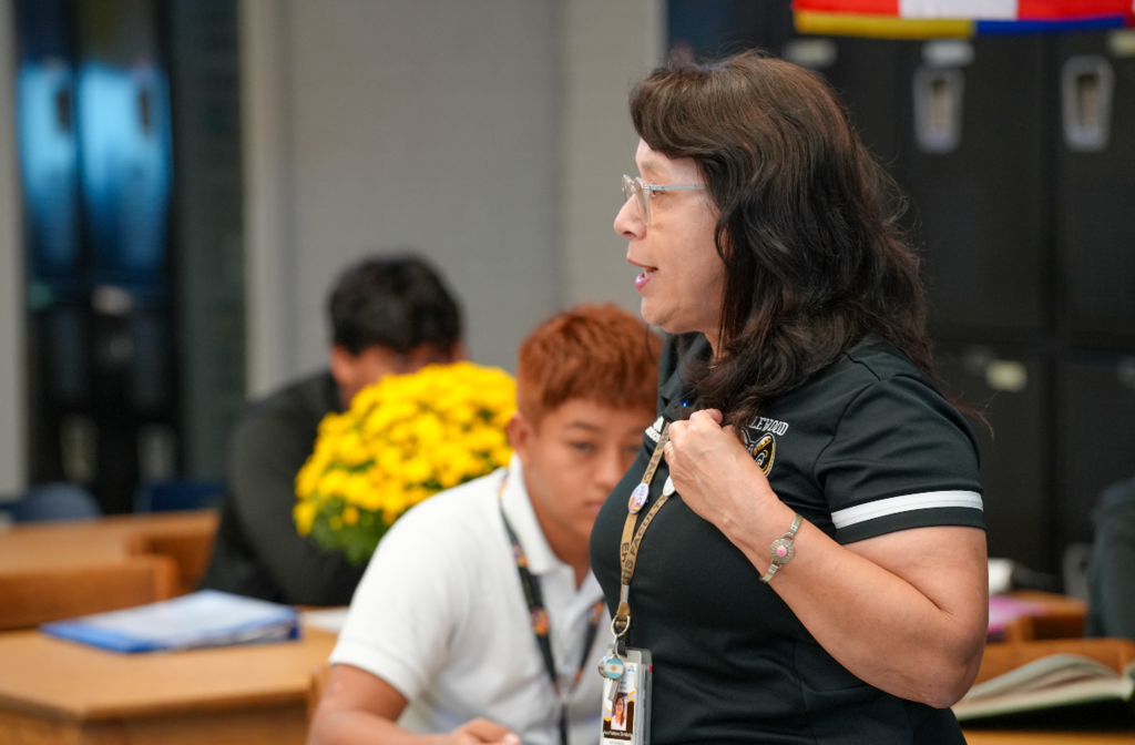 Señora Martin stands in front of black lockers, speaking to her class while wearing an Englewood High School Rams polo and ID badge.
