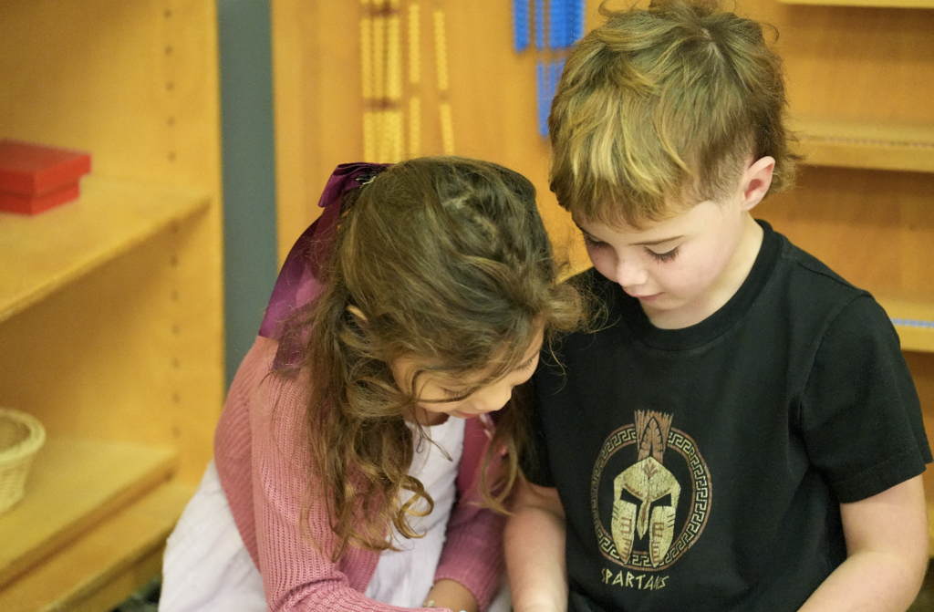 Two young students lean over a Montessori work table together, concentrating on a hands-on learning activity at J. Allen Axson Elementary.