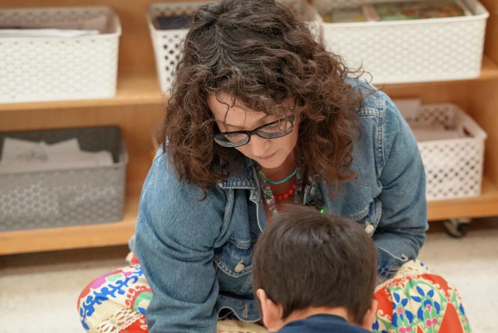 Teacher Shannon Simon sits on the classroom floor smiling while working with a student during Montessori instruction.