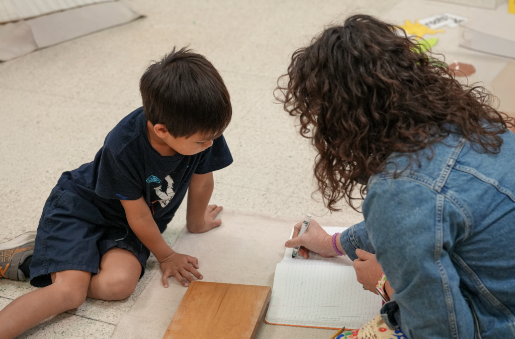 A young student carefully writes on paper while working independently at a Montessori desk in the classroom.