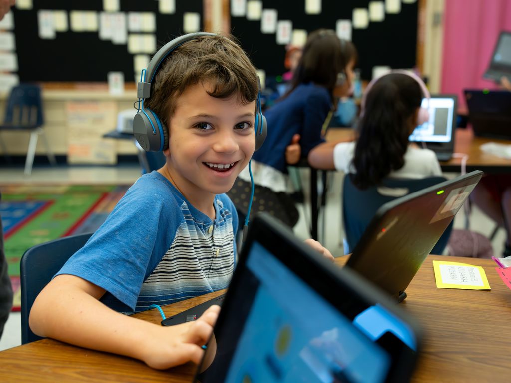 A child wearing headphones smiles at the camera as he works on his laptop in the classroom.