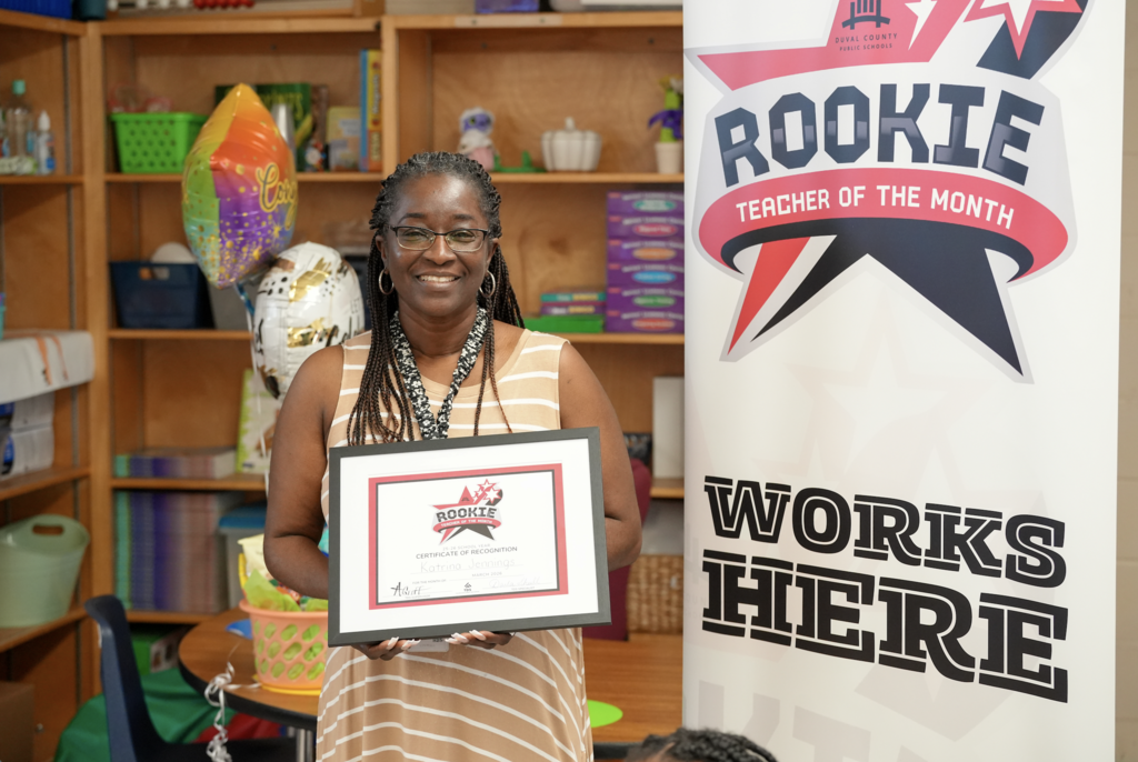 Katrina Jennings stands in a classroom holding a Rookie Teacher of the Month certificate, smiling between two colleagues next to a “Rookie Teacher of the Month Works Here” banner.