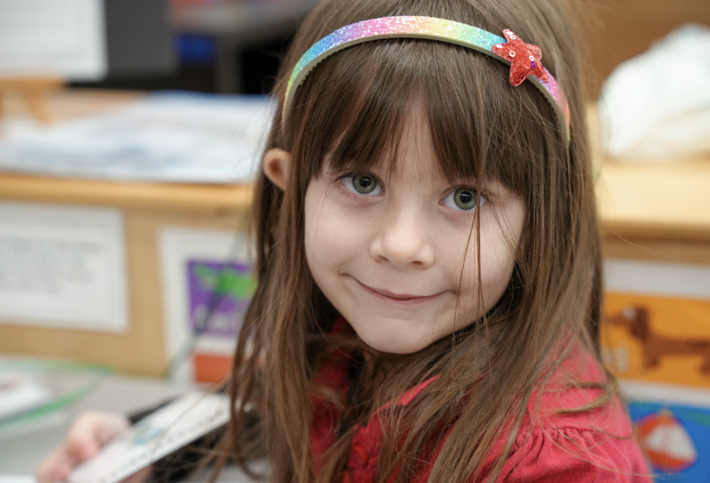 Young student smiling at the camera while sitting at a classroom table.
