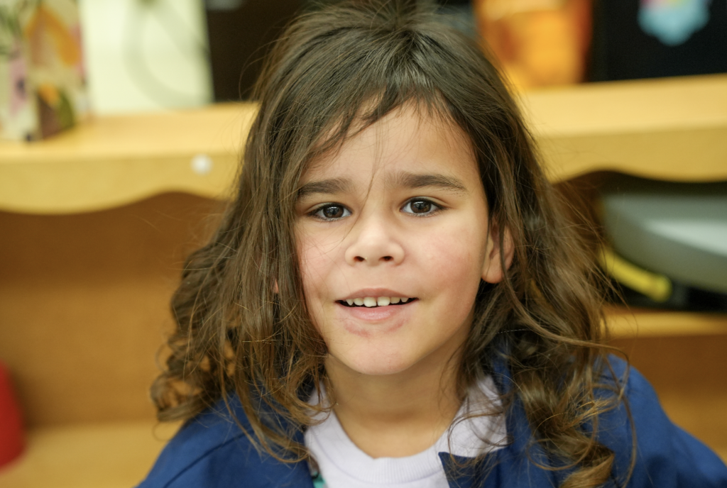 Young student smiling at the camera while sitting at a classroom table.