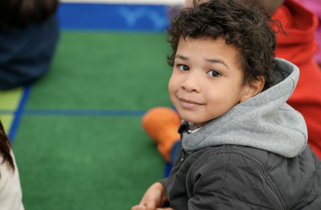Young student smiling at the camera while sitting at a classroom table.