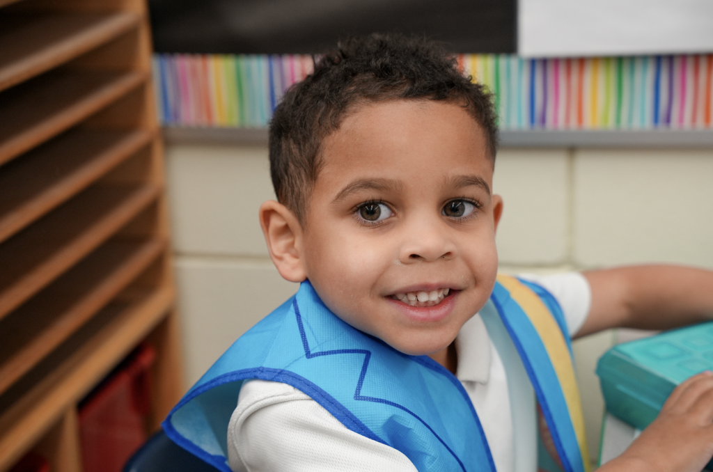 Young student smiling at the camera while sitting at a classroom table.