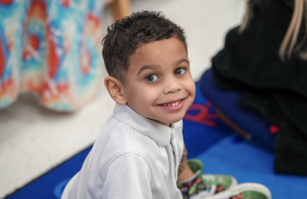 Young student smiling at the camera while sitting at a classroom table.
