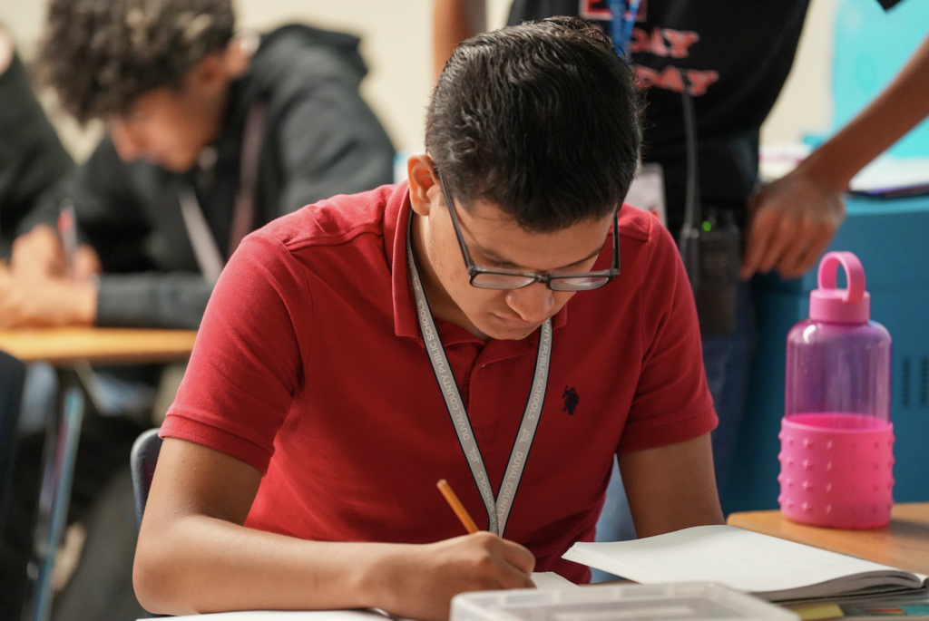 A student in a red shirt looks down and writes in a notebook during a classroom activity.