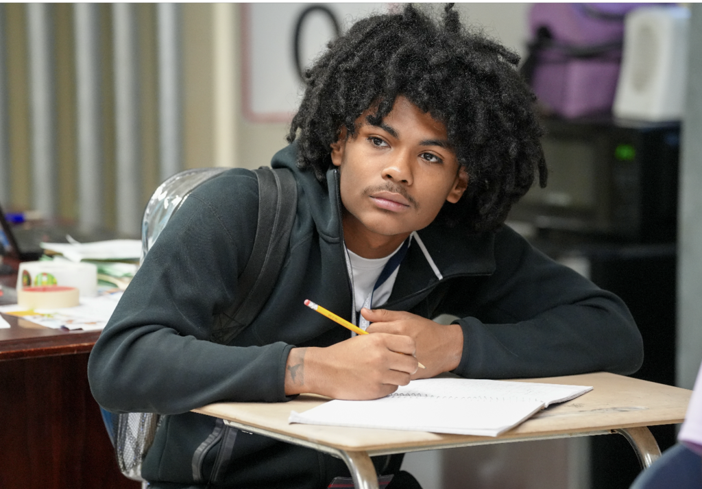 A student in a black hoodie writes in a notebook while listening during a lesson.