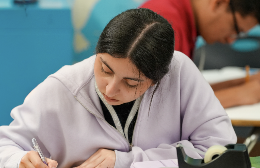 A student leans over their desk writing in a notebook during class.