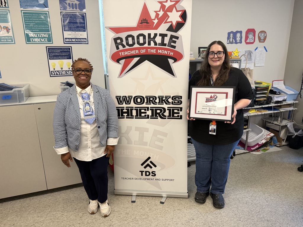 Two women stand in front of a "Rookie Teacher of the Month" banner. One holds an award.
