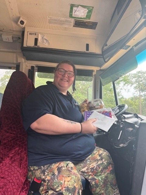 A woman sits in the driver's seat of a bus, holding a paper, and smiling.