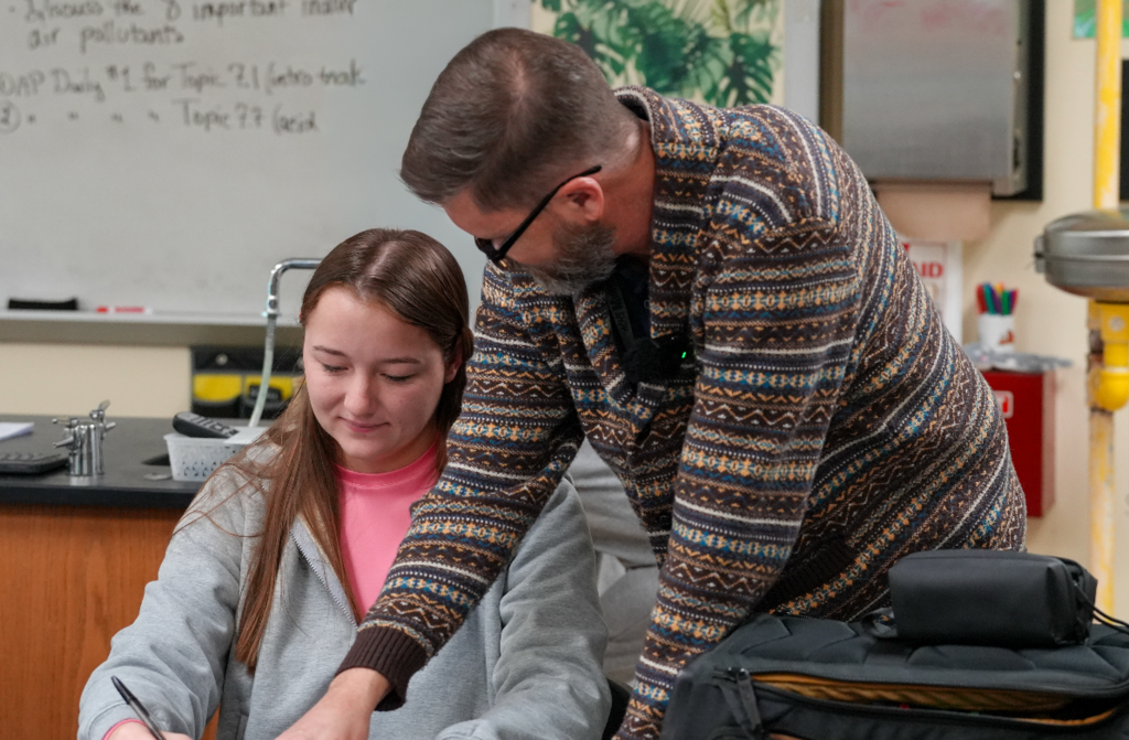 Dr. Scott Sowell, an older teacher, leans over to help a student with a classroom assignment.