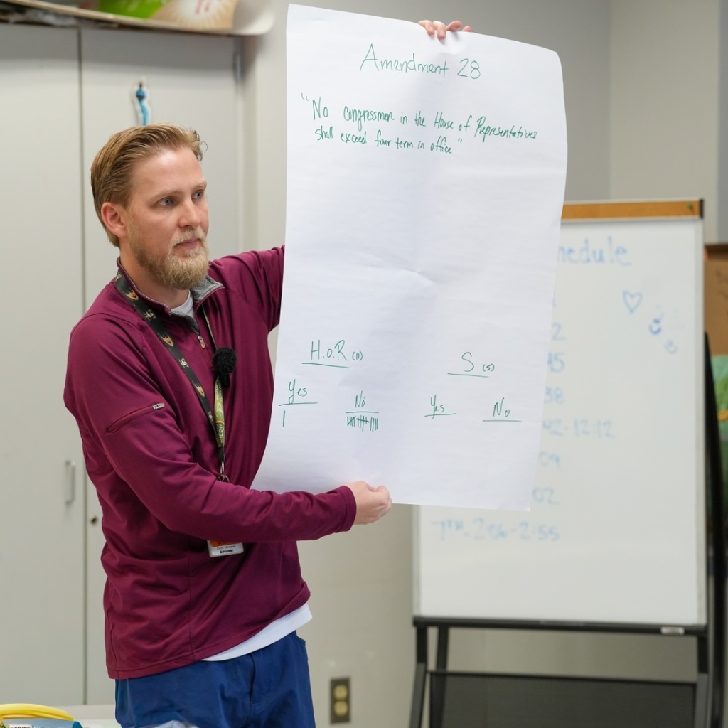 Andrew Ohmann red sweatshirt and blue pants holds a paper with written content, standing in front of a whiteboard.
