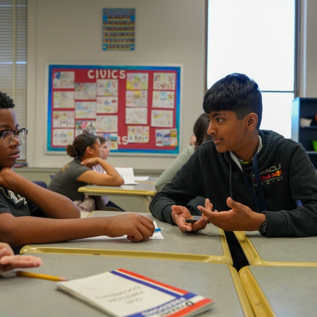 Two boys in a classroom; one is gesturing and speaking, while the other listens attentively. A Civics bulletin board is on the wall.