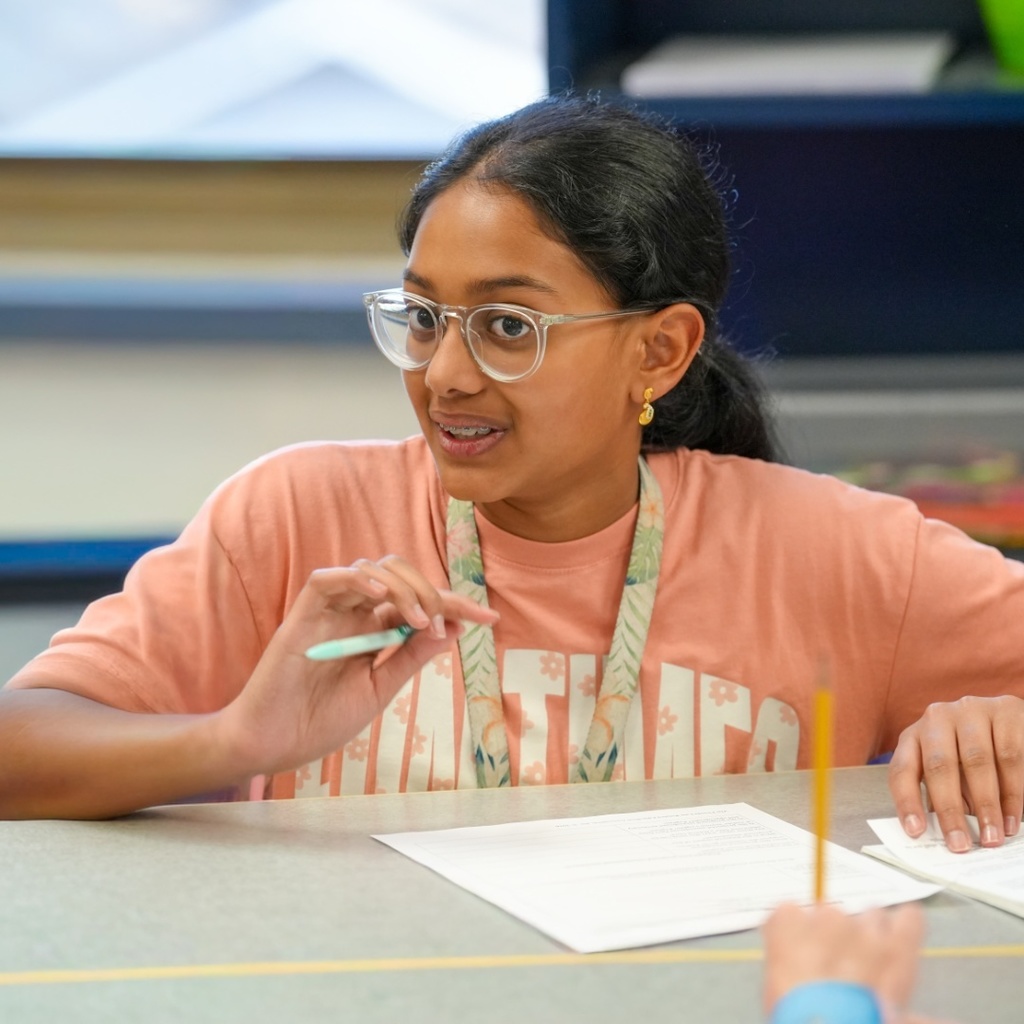 Woman with glasses seated at a table discussing something with another person. Papers and a pencil on the table.