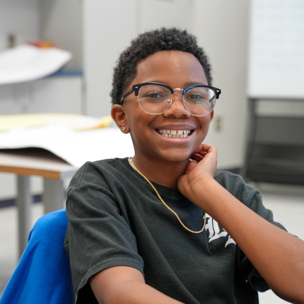 A smiling person in glasses sits in a chair in a classroom. The person wears a black T-shirt and a gold necklace.