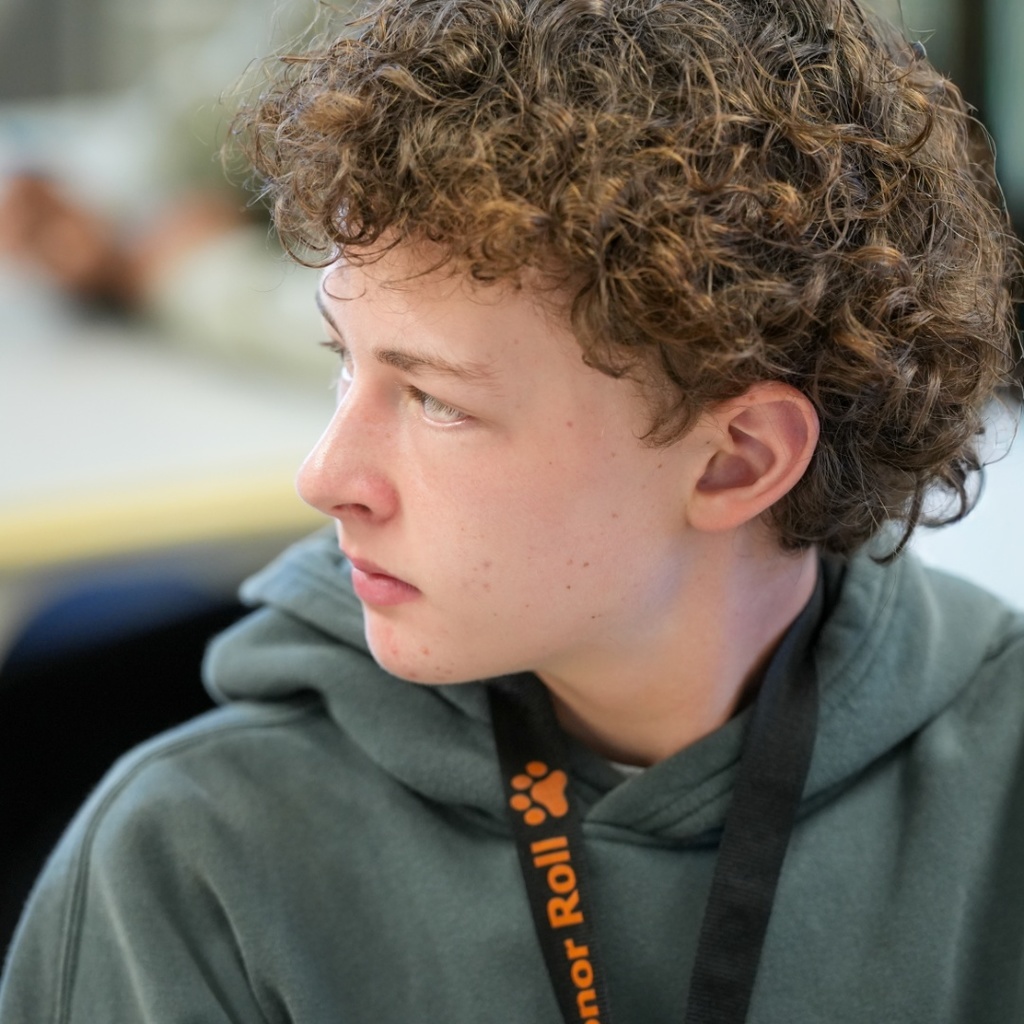 A young person with curly brown hair wears a green hoodie with a black lanyard.